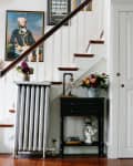Staircase with a portrait, radiator, and floral arrangements; person seated in adjacent room.