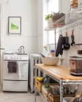 Small kitchen with wooden cabinets, white stove, metal shelves, potted plants, and colorful rug near the door.