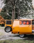 Orange camper van with open side door, revealing a small kitchen setup inside, parked in a leafy park setting.