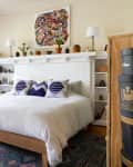 Bedroom with a white bed, blue patterned pillows, colorful artwork, and potted plants on a white brick mantel.