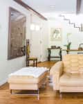 Beige armchair and ottoman in a bright living room with wooden floors, staircase, and eclectic decor.