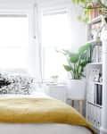 Bright bedroom with a yellow blanket, patterned pillows, large plants, and bookshelves by bay windows.