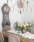 Dining room with rustic table, floral centerpiece, vintage clock, chandelier, and oval mirror above a white cabinet.