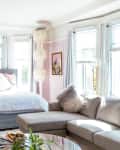 Bright bedroom with a gray bed, white bedding, pink and patterned pillows, gray sectional sofa, and large windows.
