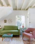 Living room with green sofa, pink chair, white rug, and wooden floor, under a staircase with vintage suitcases.