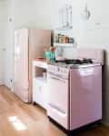 Retro kitchen with pink fridge and stove, white cabinet, wooden utensils, and vintage tins on a shelf.
