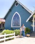 Couple with a dog standing in front of a blue house with a large arched window and garden.