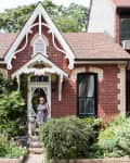 Victorian-style brick house with ornate gable, lush garden, and a person with a dog at the entrance.