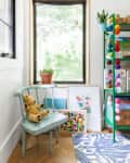 Children's room corner with a teddy bear on a mint chair, colorful art, potted plant, and a shelf with books and pom-poms.