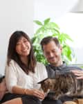 Couple sitting on a sofa with a tabby cat, surrounded by green plants and a modern floor lamp.