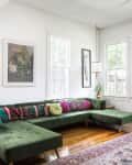 Green velvet sectional sofa with colorful pillows in a bright living room, featuring wooden floors and eclectic wall art.