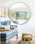 Cozy bedroom with a blue sofa, round mirror, and a bed with white and yellow bedding, featuring a beach painting.