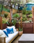 Outdoor patio with wooden steps, string lights, white sofa, blue pillows, and lush greenery.