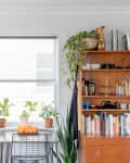 Home bar with wooden shelves, liquor bottles, glassware, plants, books, and a small table with oranges by a window.