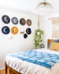 Bedroom with wooden bed, blue throw, macramé wall hanging, hat display, and potted plants.