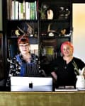 Two women standing behind a counter with bookshelves, plants, and a lamp in a cozy room.