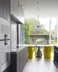 Modern kitchen with gray cabinets, marble countertops, yellow chairs, and a large window overlooking a garden.