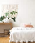 A bedroom with light wood floors, a white bed, minimal art, and a tropical green plant on a wooden table.