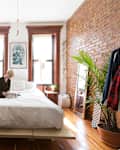 Cozy bedroom with a brick wall, plants, wooden furniture, and a person reading on a bed under large windows.
