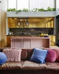 Modern living room with brown leather sofa, colorful pillows, open kitchen with gold cabinets, and large windows.