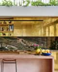 Modern kitchen with gold cabinets, marble backsplash, pink island, and plants above. Two stools and fruit bowl on the counter.