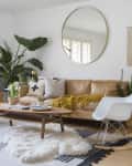 Living room with tan leather sofa, yellow throw, white chair, round mirror, plants, and macramé wall hanging.