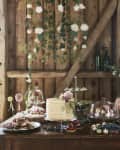 Rustic dessert table with layered cake, macarons, cupcakes, and flowers against a wooden backdrop with hanging greenery.