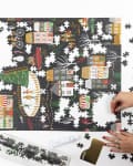 Hands assembling a colorful winter village puzzle with scattered pieces, candle, and decorative box nearby.