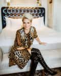 Woman in leopard print dress and black knee-high boots sitting on a bench in a stylish bedroom.