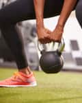 Person in gym holding a kettlebell, wearing black leggings and orange sneakers on green turf.