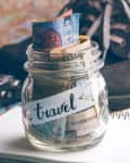Jar labeled "travel" filled with various currencies, next to a vintage camera, notebook, and pencil on a map.