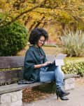 Woman sitting on a park bench using a laptop, surrounded by autumn foliage.