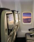 Airplane cabin with three empty seats, seatback screens, and sunset view through windows.