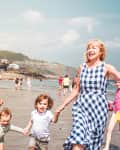 Two women and two children holding hands and running on a crowded beach, with hills and houses in the background.
