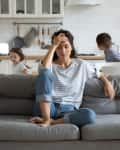 Woman sitting on a gray sofa in a kitchen, looking stressed, with two children playing in the background.