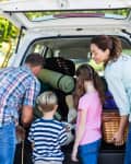 Family packing a car trunk with camping gear, including a rolled-up mat and picnic basket, under sunny trees.