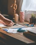 Person crafting at a wooden desk with scissors, twine, potted plant, and art supplies near a window.