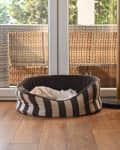 Striped pet bed on wooden floor near glass doors with wicker furniture outside.