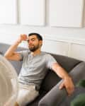 Man sitting on a gray sofa, cooling off with a fan, holding a cold drink, in a bright living room with a floor lamp.