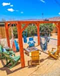 Backyard with wooden pergola, colorful chairs, hammocks, fire pit, and shaded dining area against a desert landscape.