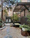 Narrow garden pathway with gravel, plants, and a seating area under a wooden pergola.