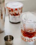Prepared cocktail on surface with shaker and jar of jam in background.