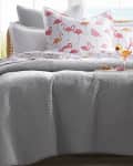 Gray quilted bedding with flamingo print pillows, wicker side table, orange lamp, and a cocktail glass.