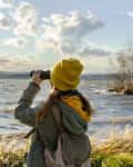 Person in yellow beanie and green jacket using binoculars by a lake under a cloudy sky.