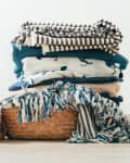 Dog lying on back next to a stack of folded blankets in various patterns and colors on a woven basket.