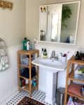 Bathroom with pedestal sink, wooden shelves, toiletries, and a mirror. Green towel on a ring holder and a potted plant visible.