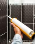 Person applying white caulk to black tiled bathroom wall with a caulking gun.