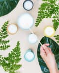 Hand lighting candles surrounded by green leaves on a beige surface.