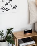 Bedside table with digital clock, potted plant, books, and beige bedding in a minimalist bedroom.
