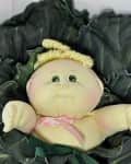 Cabbage Patch doll with green eyes and blonde hair nestled in large green leaves.